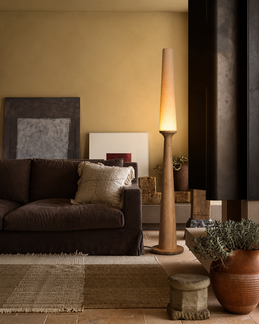 A tall, modern floor lamp with a tapered wooden shade and a warm glowing light, standing in a cozy living room next to a dark brown sofa and textured rugs.