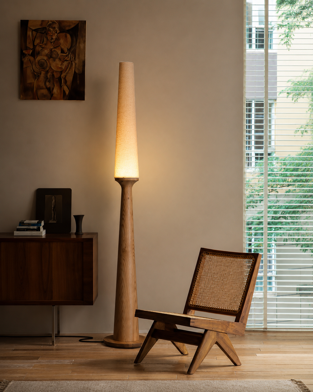 A tall floor lamp with a tapered beige shade and warm glowing light stands beside a wooden chair with a woven cane back in a modern, minimalist room with a window and blinds.