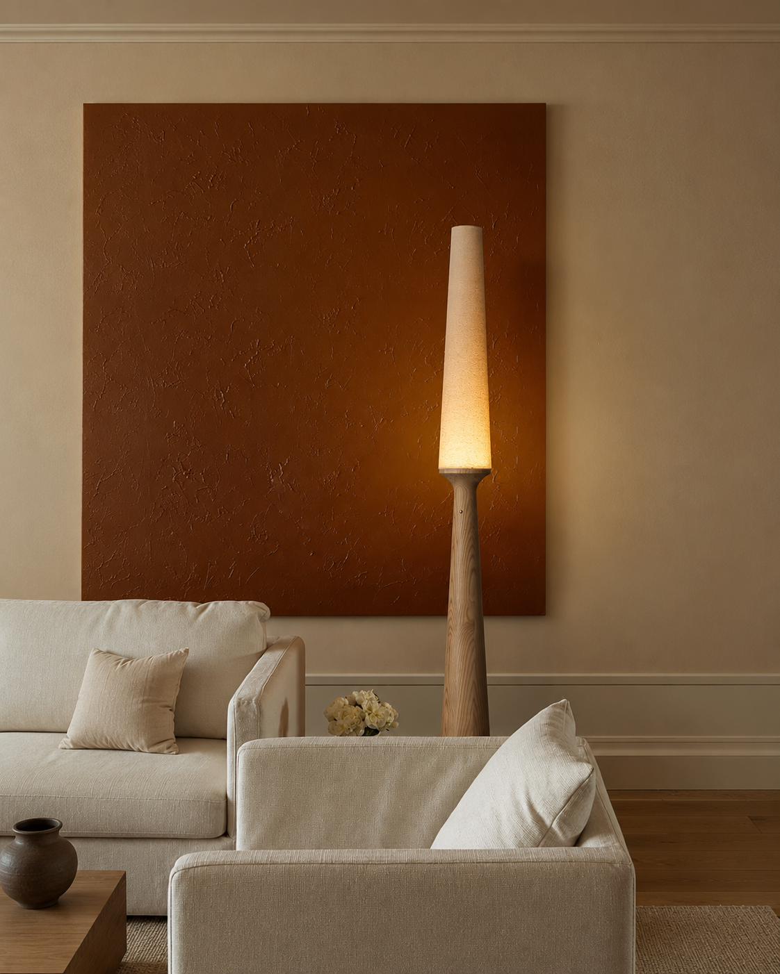 A modern living room scene featuring a tall floor lamp with a tapered white shade and a warm glowing light, positioned next to a large textured brown wall art piece above a cream-colored sofa.