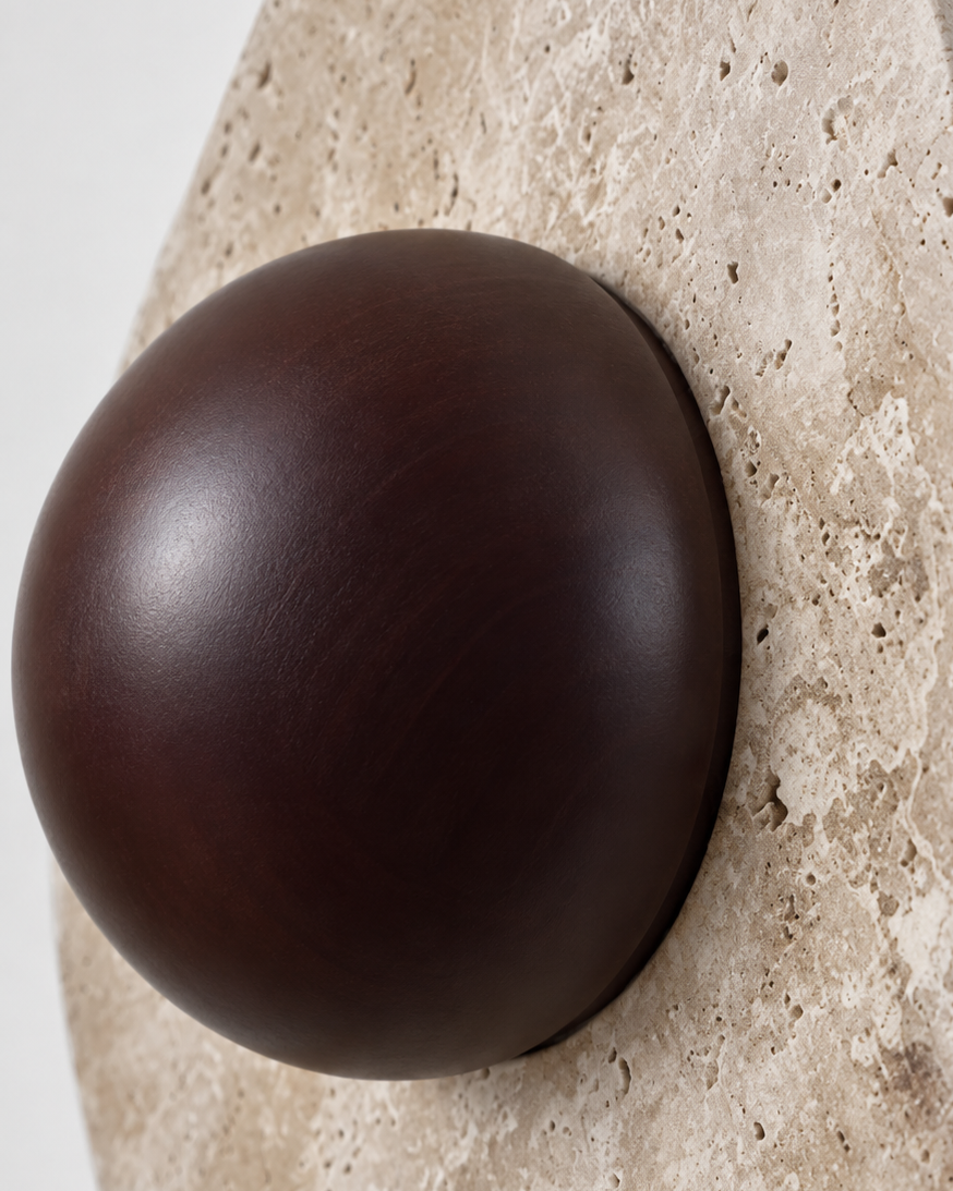 Close-up of a wall-mounted sculptural light fixture featuring a dark brown spherical shade set into a beige travertine stone disc.