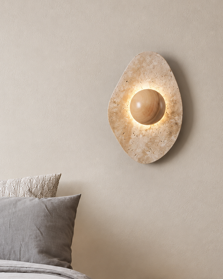 A modern oval wall sconce with a warm glowing central bulb, mounted on a textured beige plaster wall above a bed with gray bedding and a small wooden side table.
